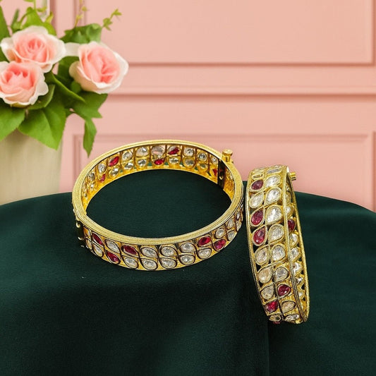 Gold - Plated Red & White Kundan Stone Bangles - Auraa Trends - Bangles -  - Auraa Trends - Pair of gold - plated bangles with alternating red and white teardrop - shaped Kundan stones, displayed on dark green velvet cloth with pink roses in the background – traditional bridal kada set.