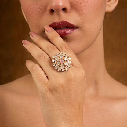 Gold-finish round cocktail ring with a blush-pink oval centre stone, clear oval stones, and American diamond halo