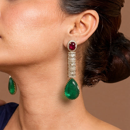 Silver-tone American diamond long drop earrings with ruby-red oval top stones, clear baguette stone rows and large emerald-green pear-cut drops on dark green velvet