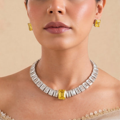Rhodium American diamond tennis necklace with emerald-cut clear stones, yellow centre stone and matching yellow stud earrings on pink background