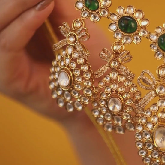 Gold-toned necklace with a braided cord and adjustable tassel tie, featuring intricate white stone detailing with oval green gemstones and multiple dangling teardrop motifs, paired with matching drop earrings, displayed on a plain light background.