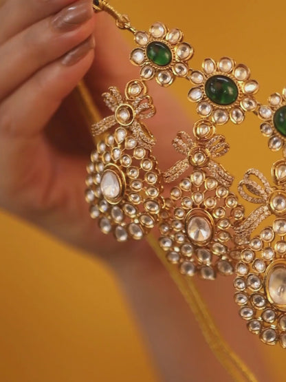 Gold-toned necklace with a braided cord and adjustable tassel tie, featuring intricate white stone detailing with oval green gemstones and multiple dangling teardrop motifs, paired with matching drop earrings, displayed on a plain light background.