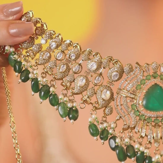 Detailed gold-toned statement necklace with intricate white stone embellishments and a prominent green teardrop centerpiece, finished with dangling green beads and pearls, paired with matching ornate drop earrings, displayed on a plain light background.